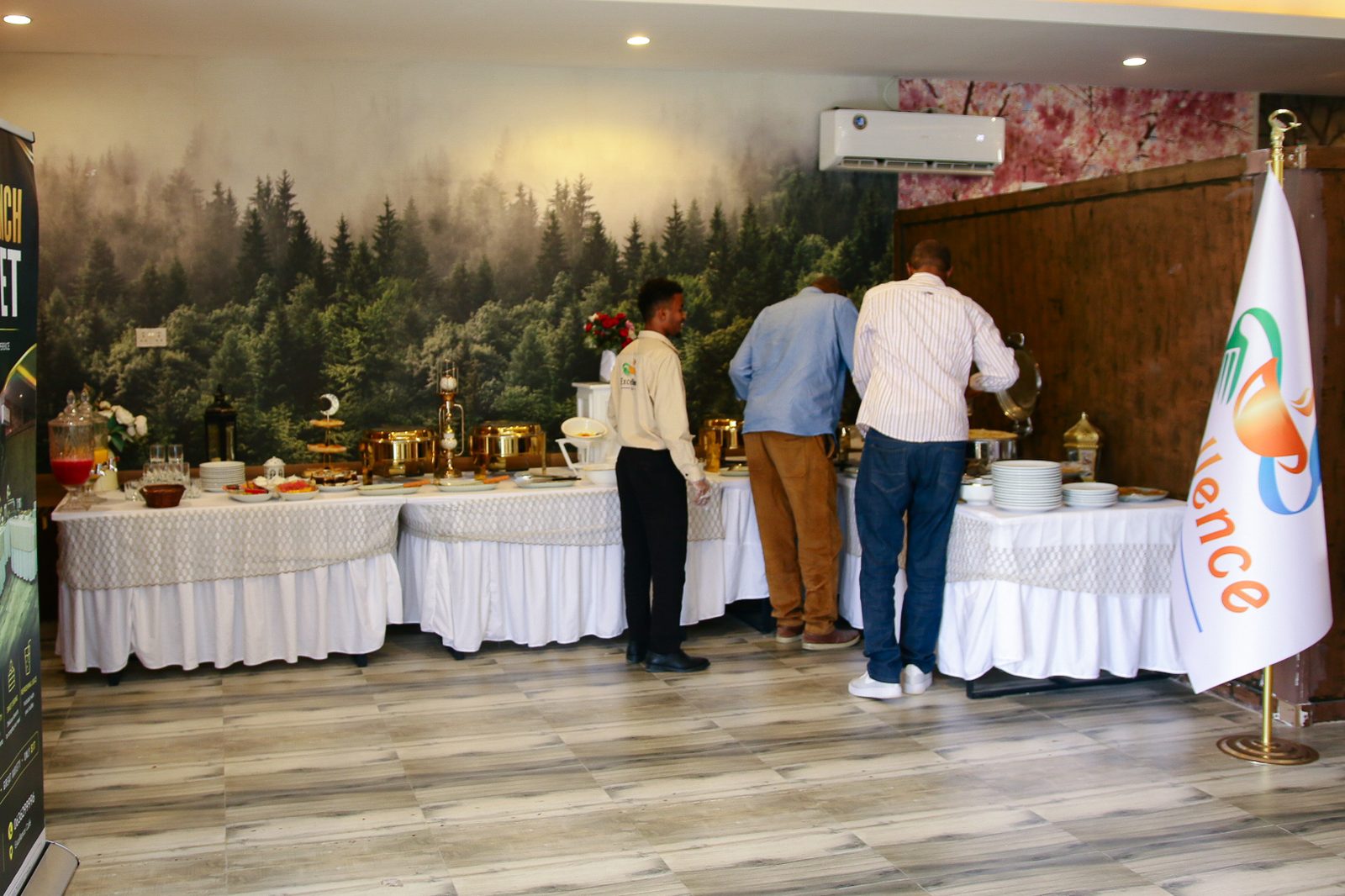 Excellence Café's indoor buffet hall — skirted white-linen tables with gold and steel chafing dishes against a forest-mural wall, staff in branded shirts.