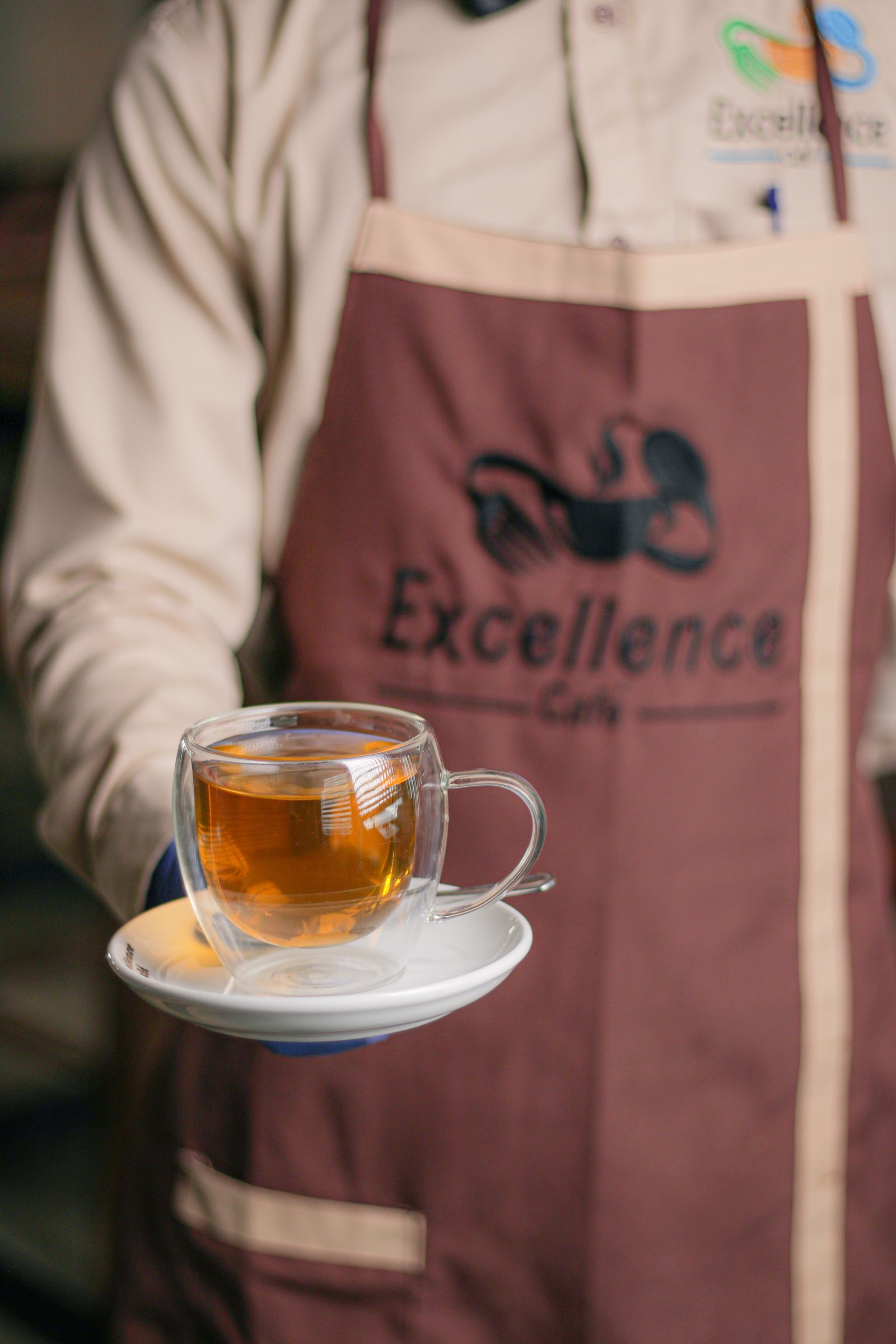 An Excellence Café staff member in a branded apron carrying a glass of tea on a saucer.