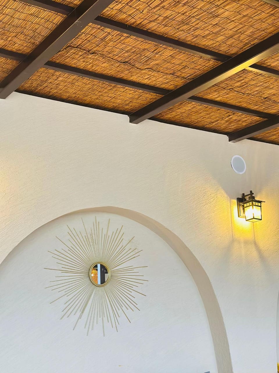 The Village Hargeisa interior — a gold sunburst mirror centred in a white plastered arch, with a lit iron wall lantern to the right, below a timber-beamed reed ceiling.