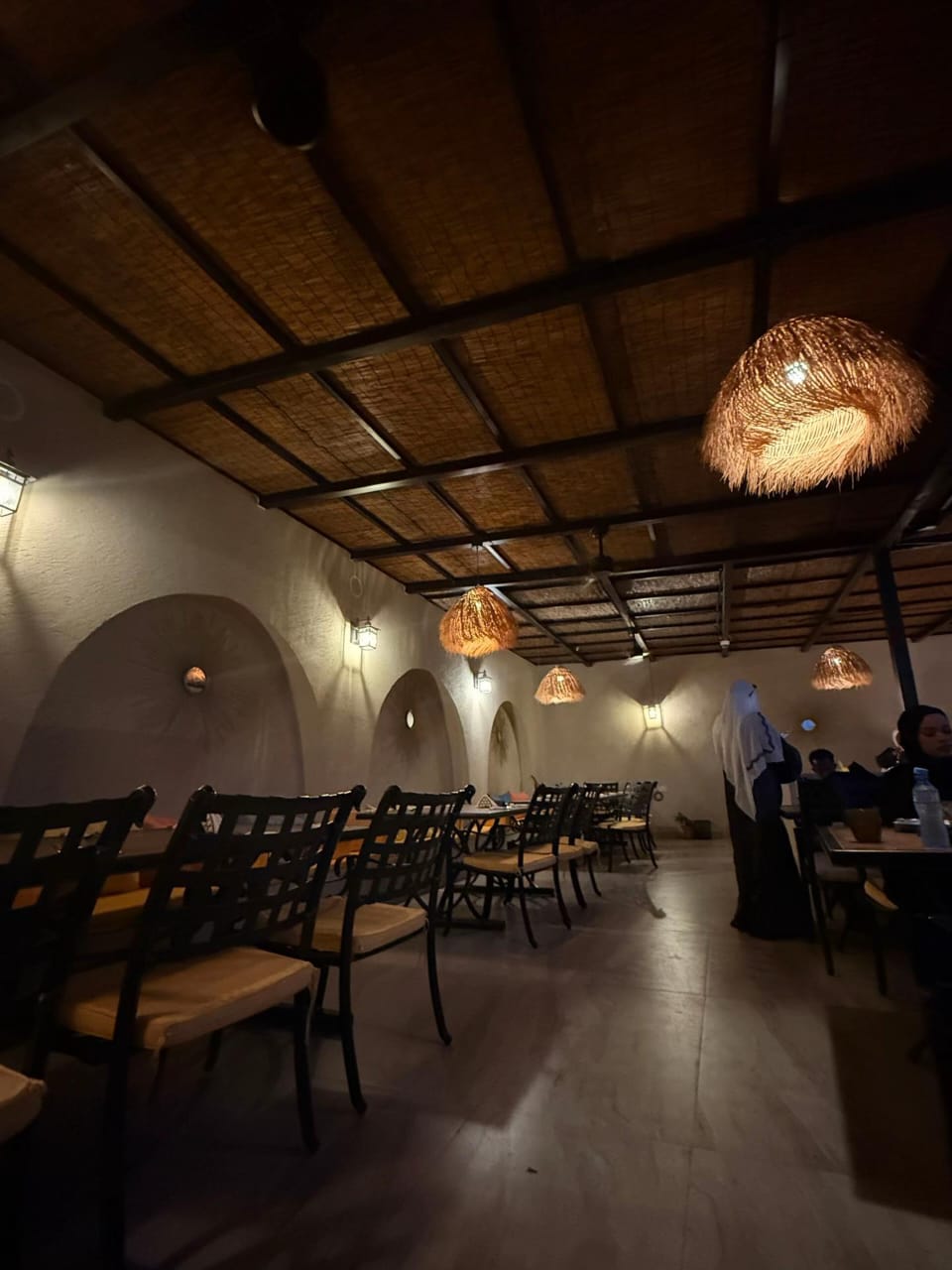 The Village Hargeisa dining hall at night — a row of woven straw pendant lamps glowing amber over iron chairs and tables, with lit arched wall niches.