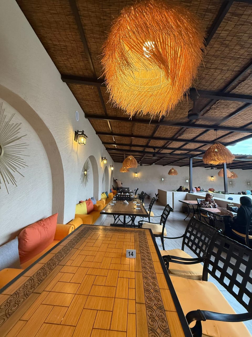The Village Hargeisa covered dining terrace — cushioned benches along a sculpted arched wall, inlaid wooden tables and iron chairs beneath a reed ceiling and a large woven pendant lamp.