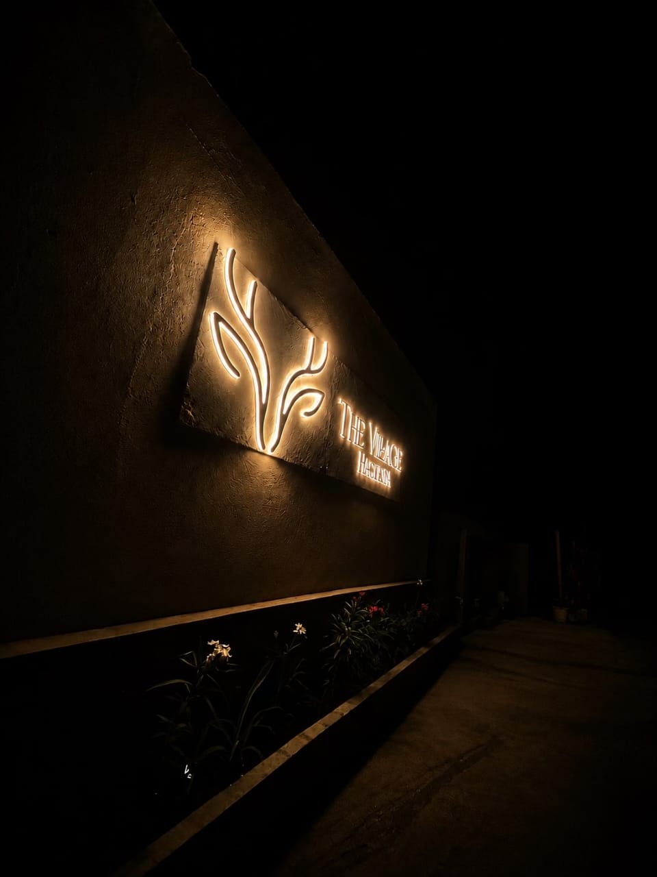 The Village Hargeisa entrance at night — the backlit restaurant emblem glowing warm on a dark textured wall above a lit flower bed.