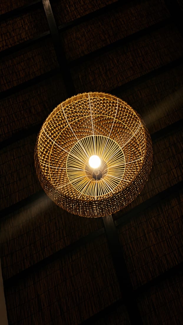 A hand-woven cane-and-rattan pendant lamp at The Village Hargeisa, lit from within against a dark reed-panelled ceiling.